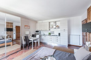 A modern kitchen with white cabinets, a microwave, and a stove. There is a dining area with a table and chairs, and a bedroom with a bed and a mirror. - Residence Albatros A1 by Wonderful Italy (Toscolano Maderno)