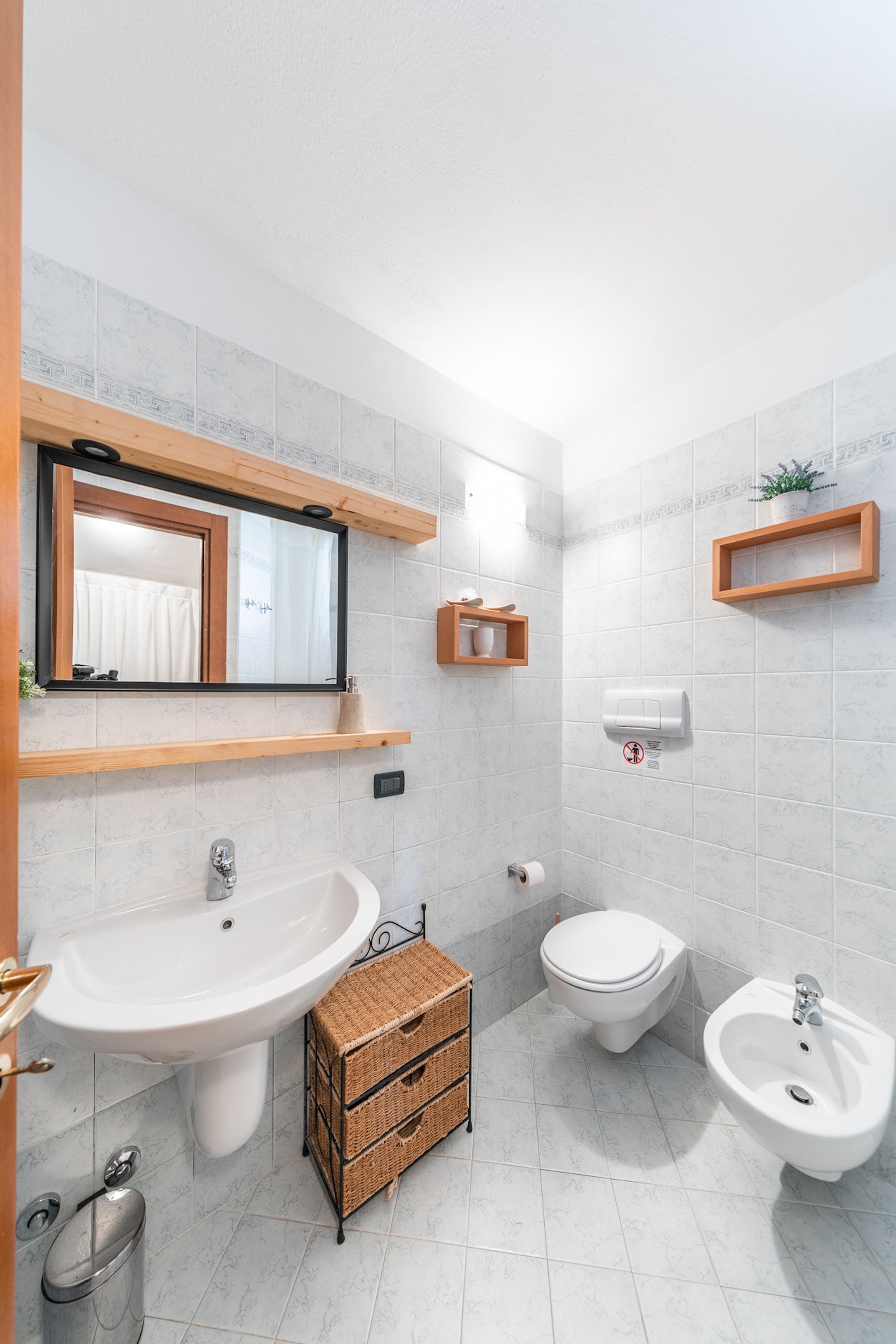 A modern bathroom with a sink, bidet, and bidet seat, wooden shelves, and a trash bin.