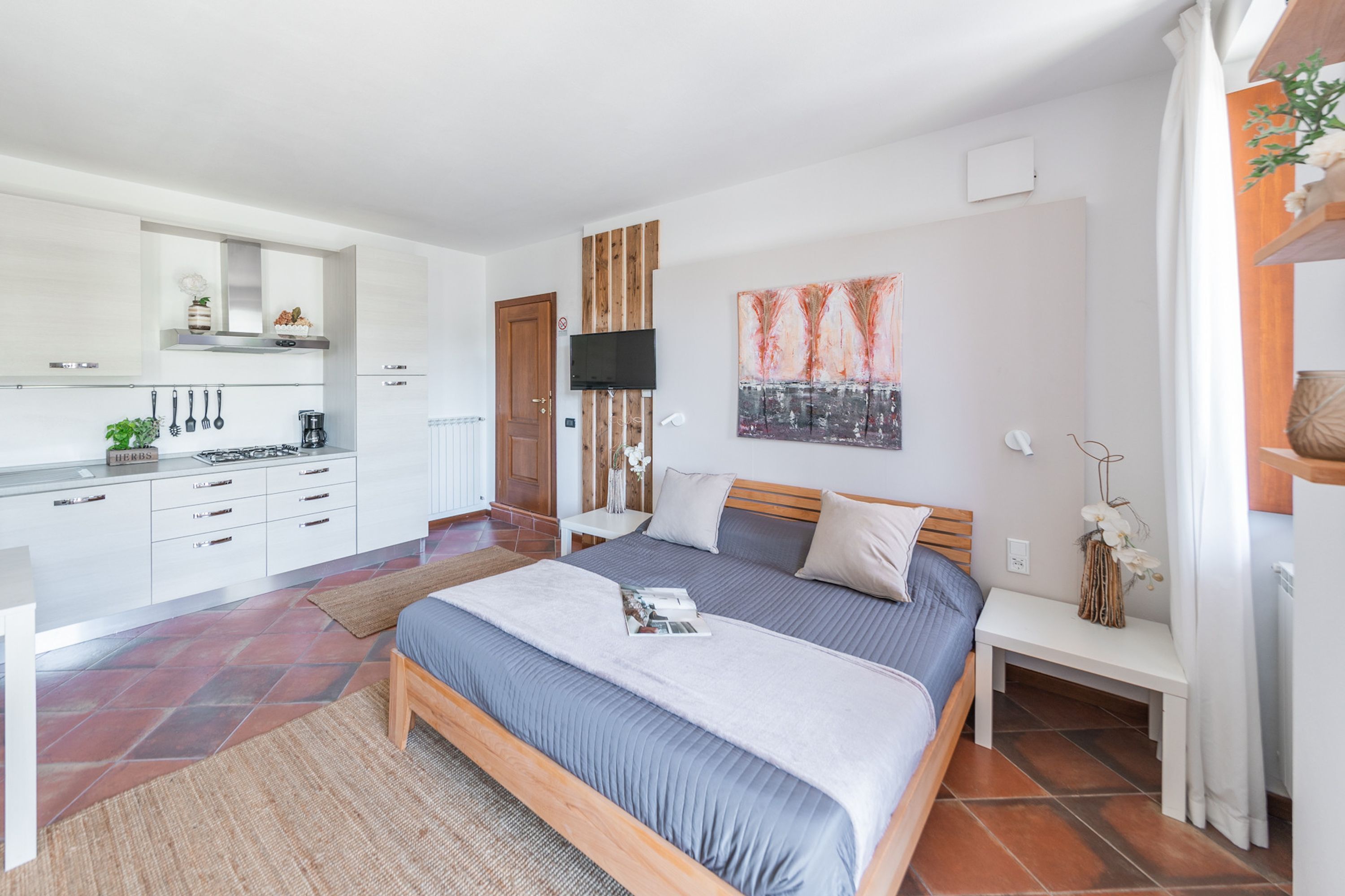 A modern bedroom with a large bed, a bedside table, a TV, and a kitchen area with cabinets and a stove.