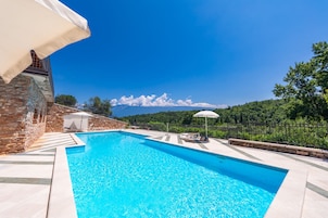 A swimming pool with a stone wall and a white umbrella, surrounded by a stone fence and greenery.