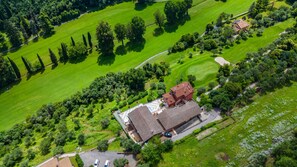 Eine Vogelperspektive zeigt ein Anwesen mit einem zentralen Gebäude, umgebenden grünen Feldern und einem Golfplatz.
