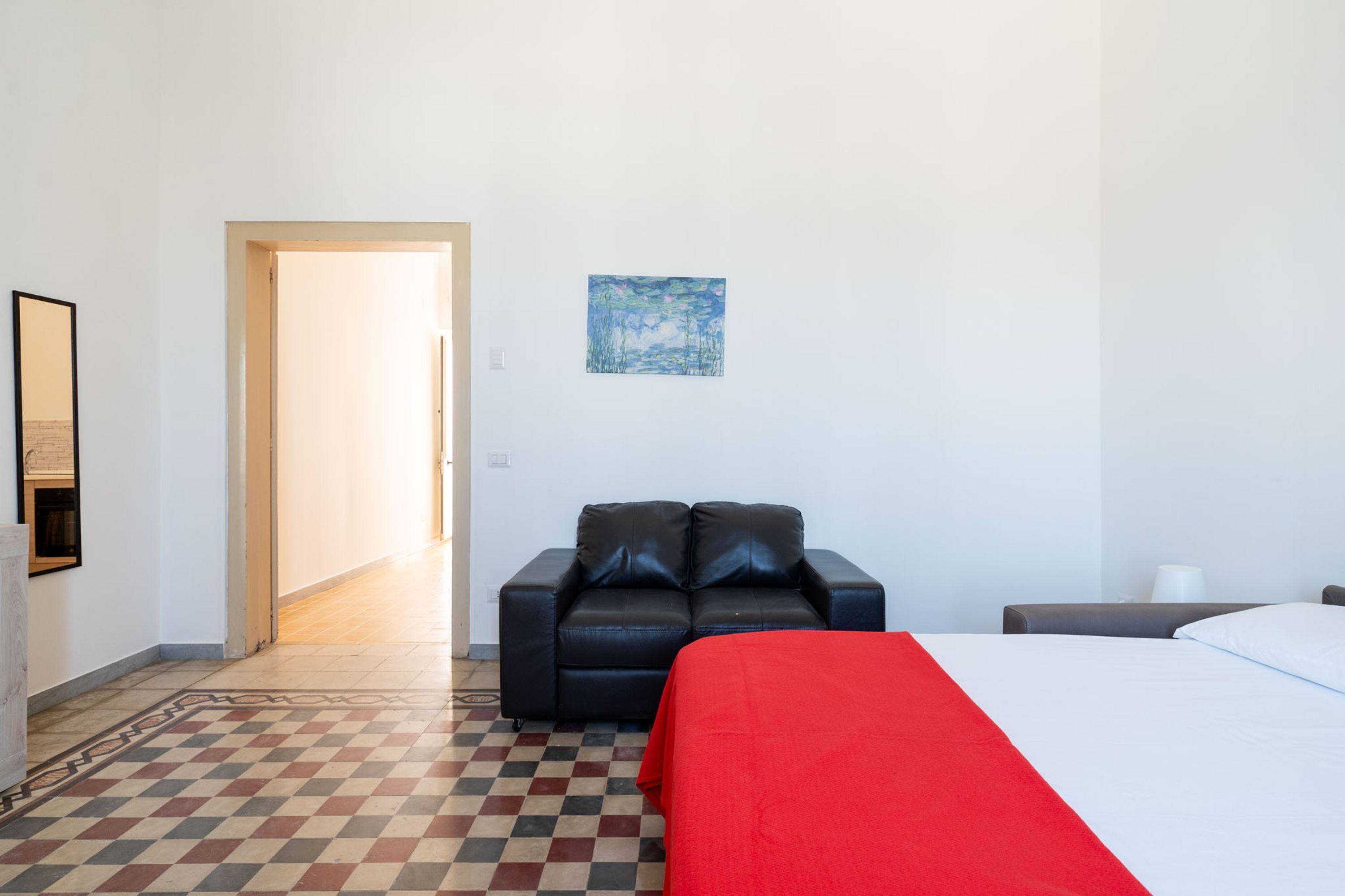 A hotel room with a checkered floor, a bed with a red blanket, a black leather sofa, and a painting on the wall.