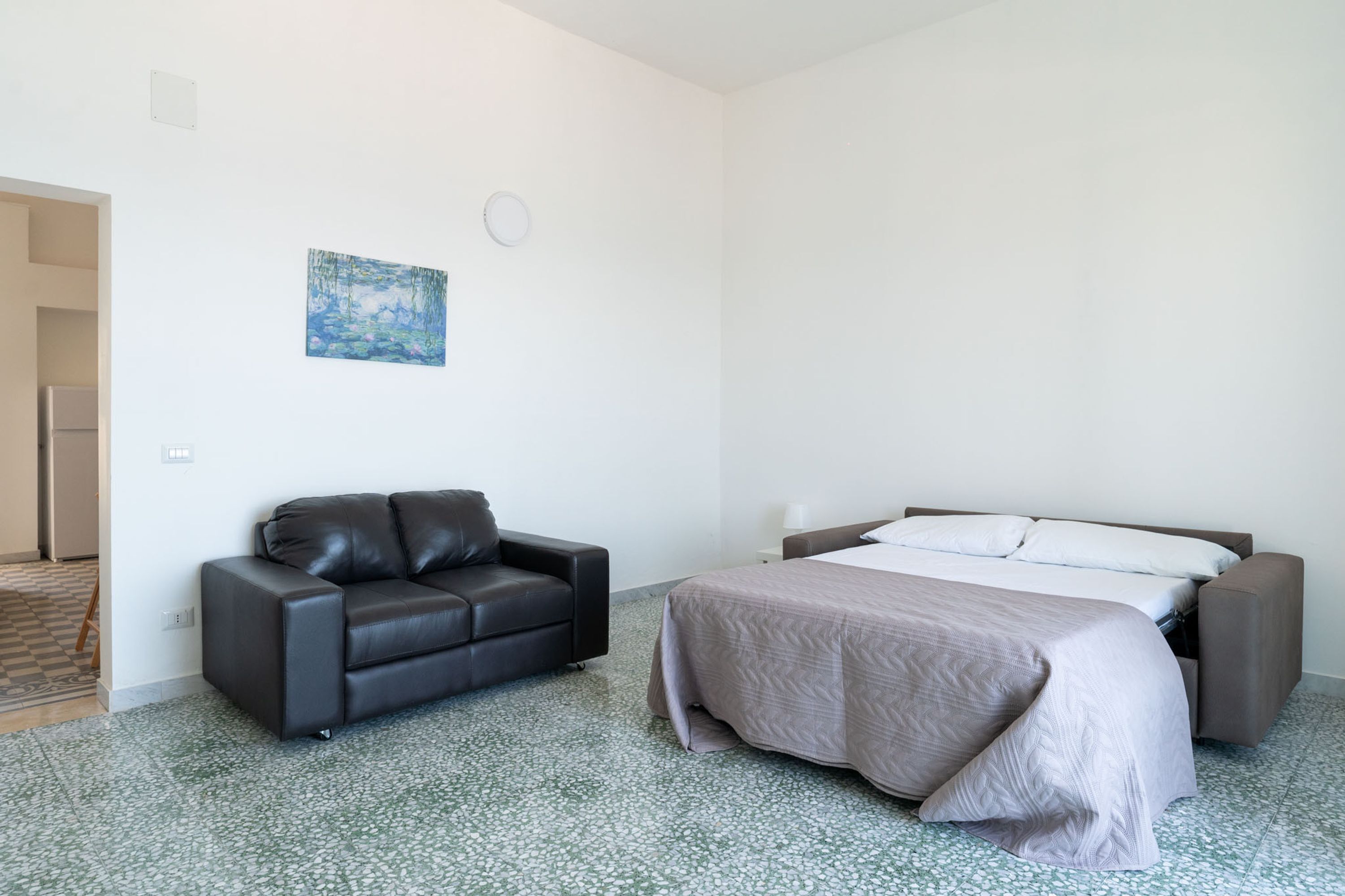 A room with a bed, a sofa, a painting on the wall, and a door leading to another room.