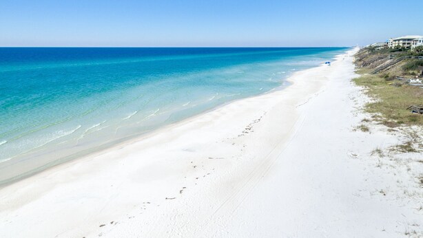 En strand med kristallklart, turkosfärgat vatten och vit sand, några byggnader i bakgrunden och en klar himmel.