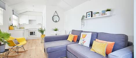 A modern living room with a grey sectional sofa, yellow and orange cushions, a dining area with a yellow chair, and a kitchen in the background.