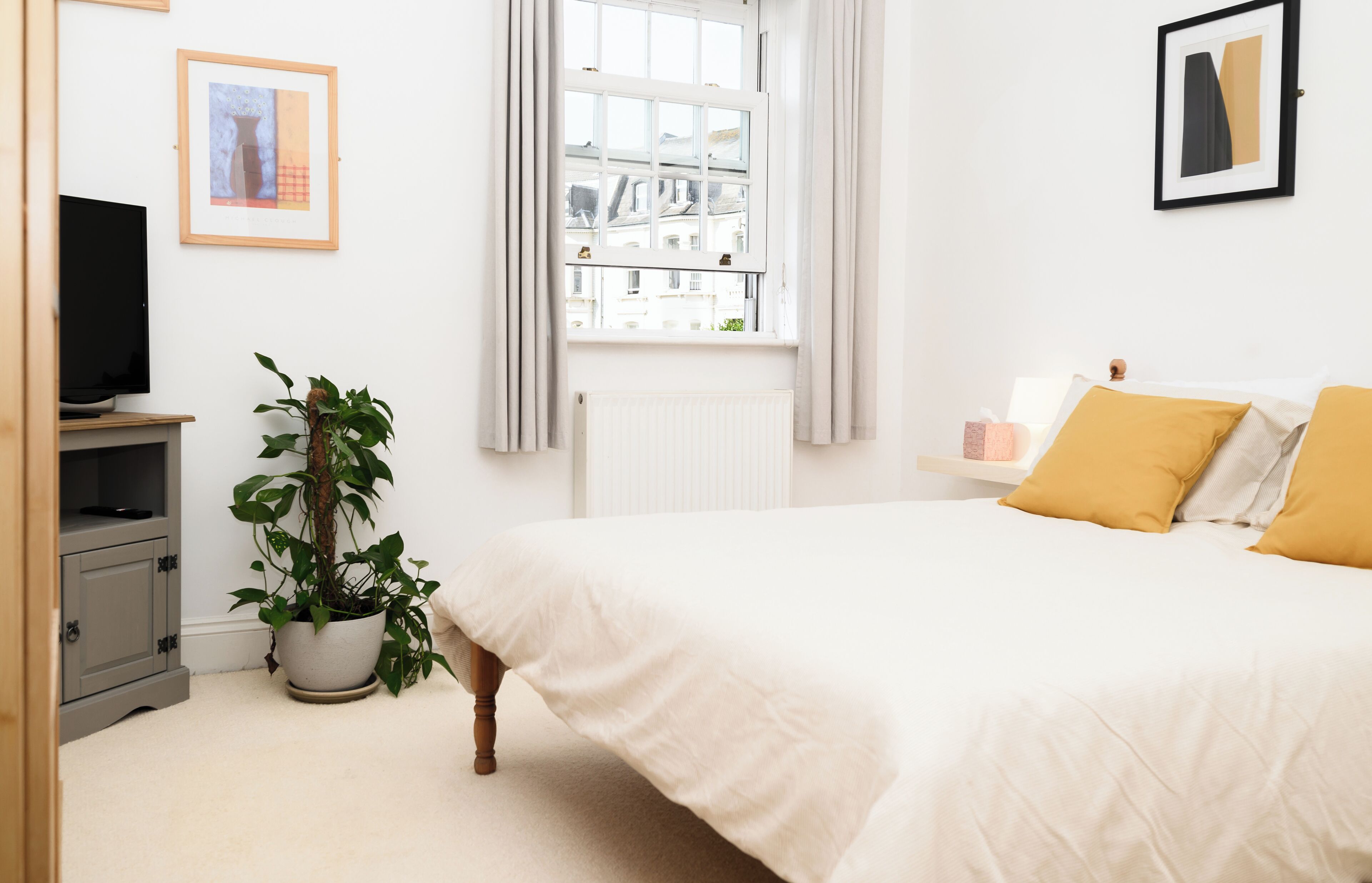 A bedroom with a bed, a potted plant, a radiator, a television, and framed artwork on the wall.