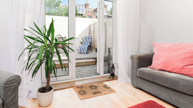 Ein Wohnzimmer mit grauem Sofa, einem roten Teppich, einer Zimmerpflanze und einer Glasschiebetür, die auf eine Terrasse führt.