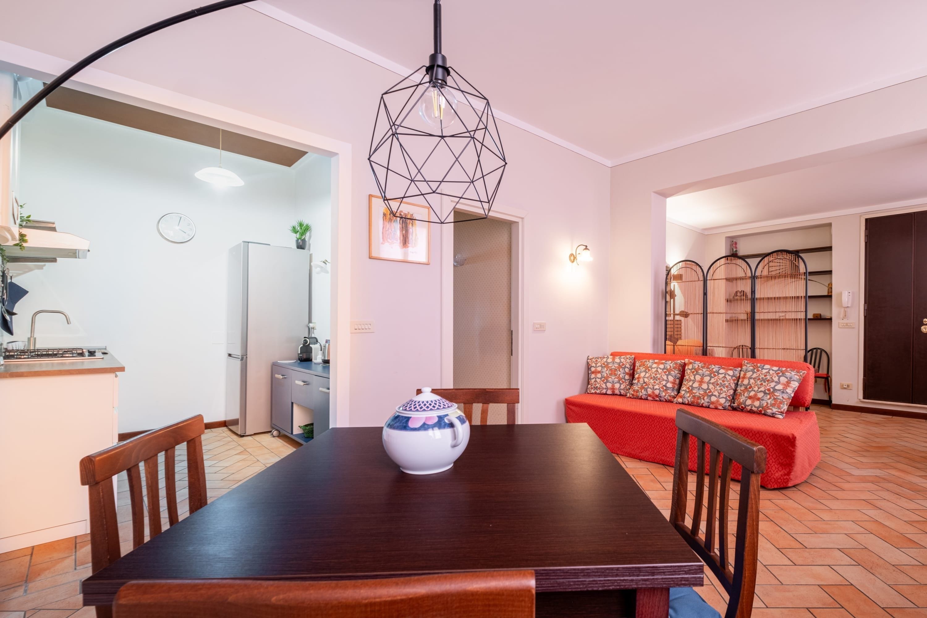 Een eethoek met een houten tafel, stoelen, een decoratieve vaas, een rode bank met opvallende kussens en een geometrische hanglamp.