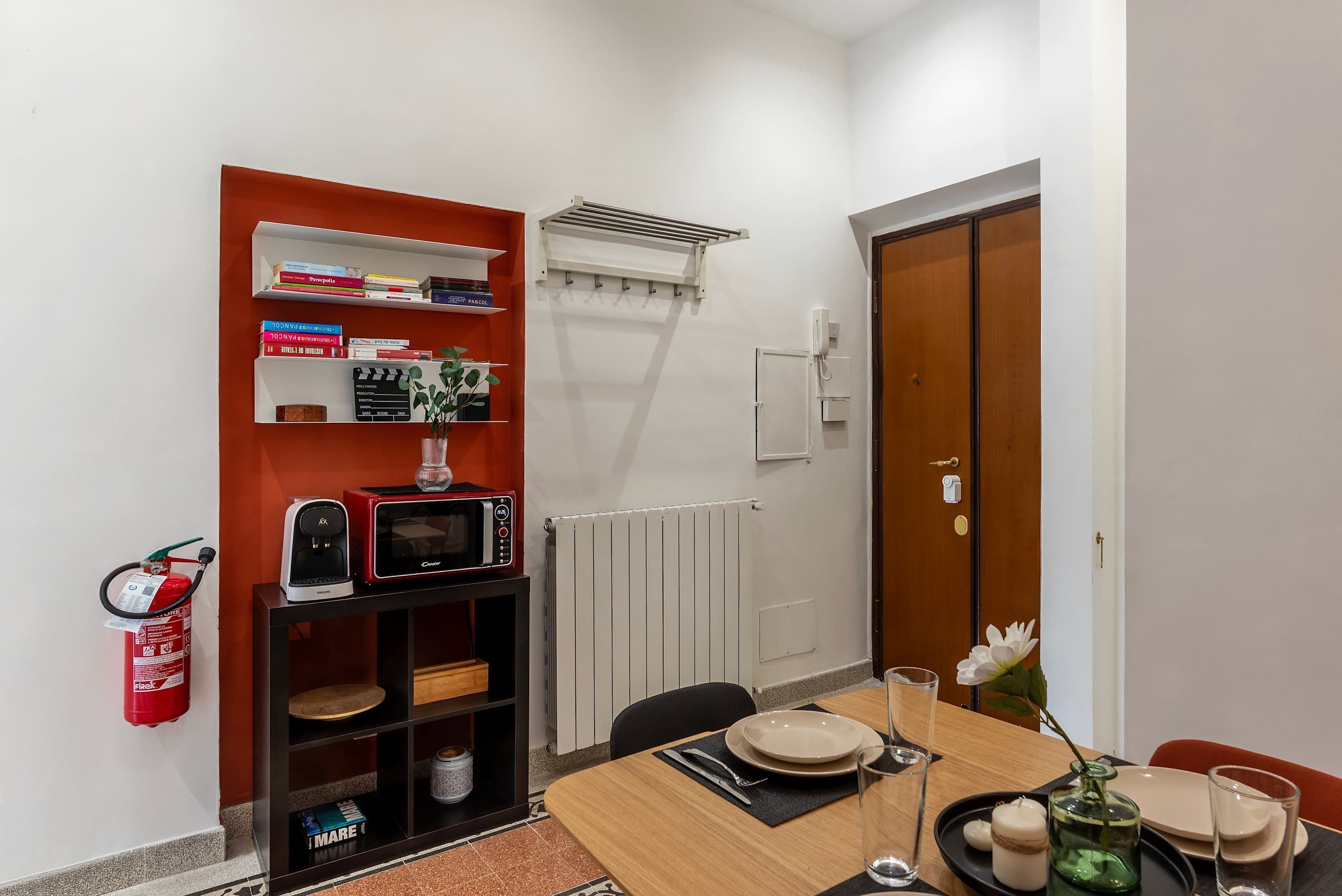 A compact dining area with a table set for two, a bookshelf with books, a microwave, and a fire extinguisher.