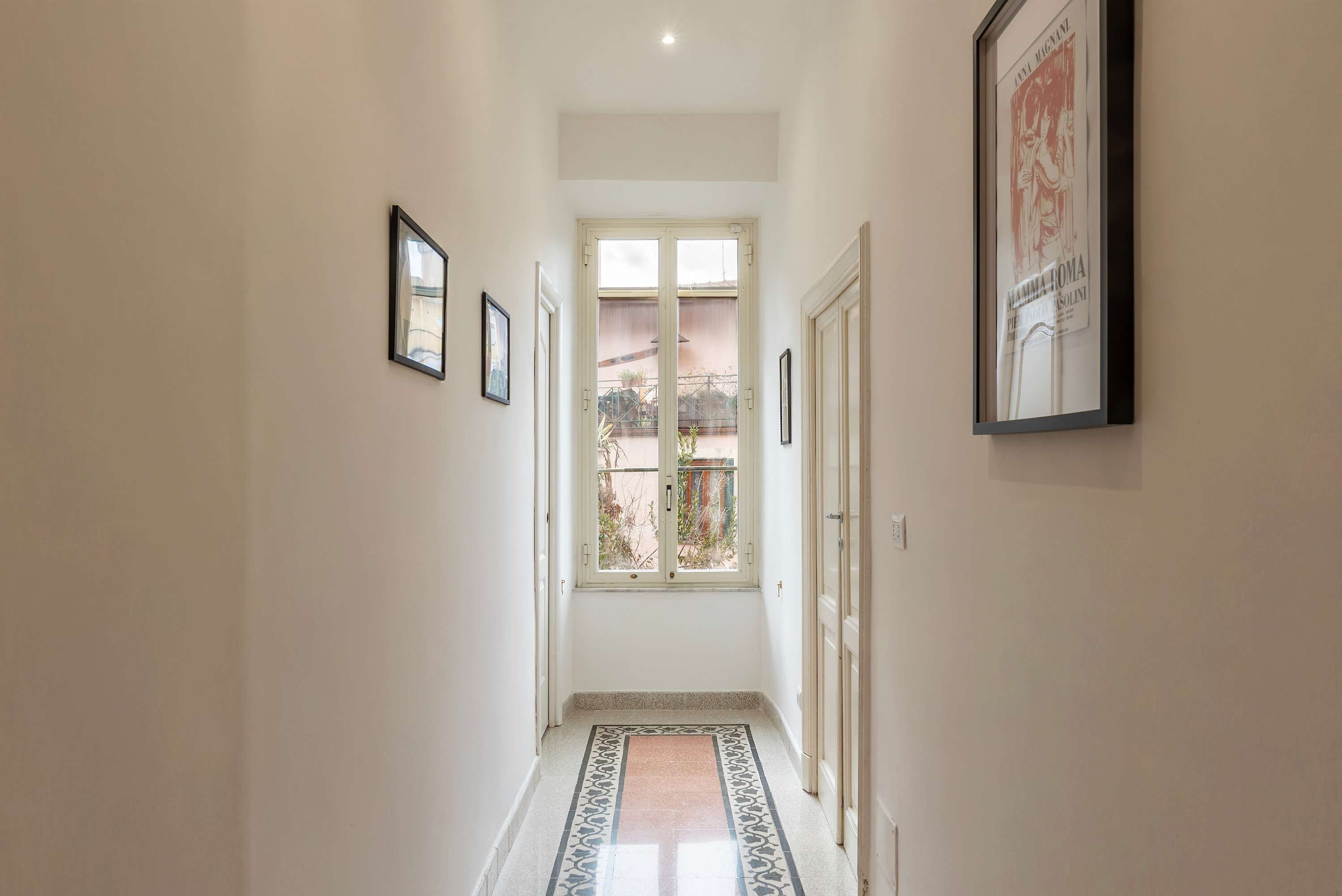 A hallway with a patterned floor, framed pictures on the walls, and a window at the end.