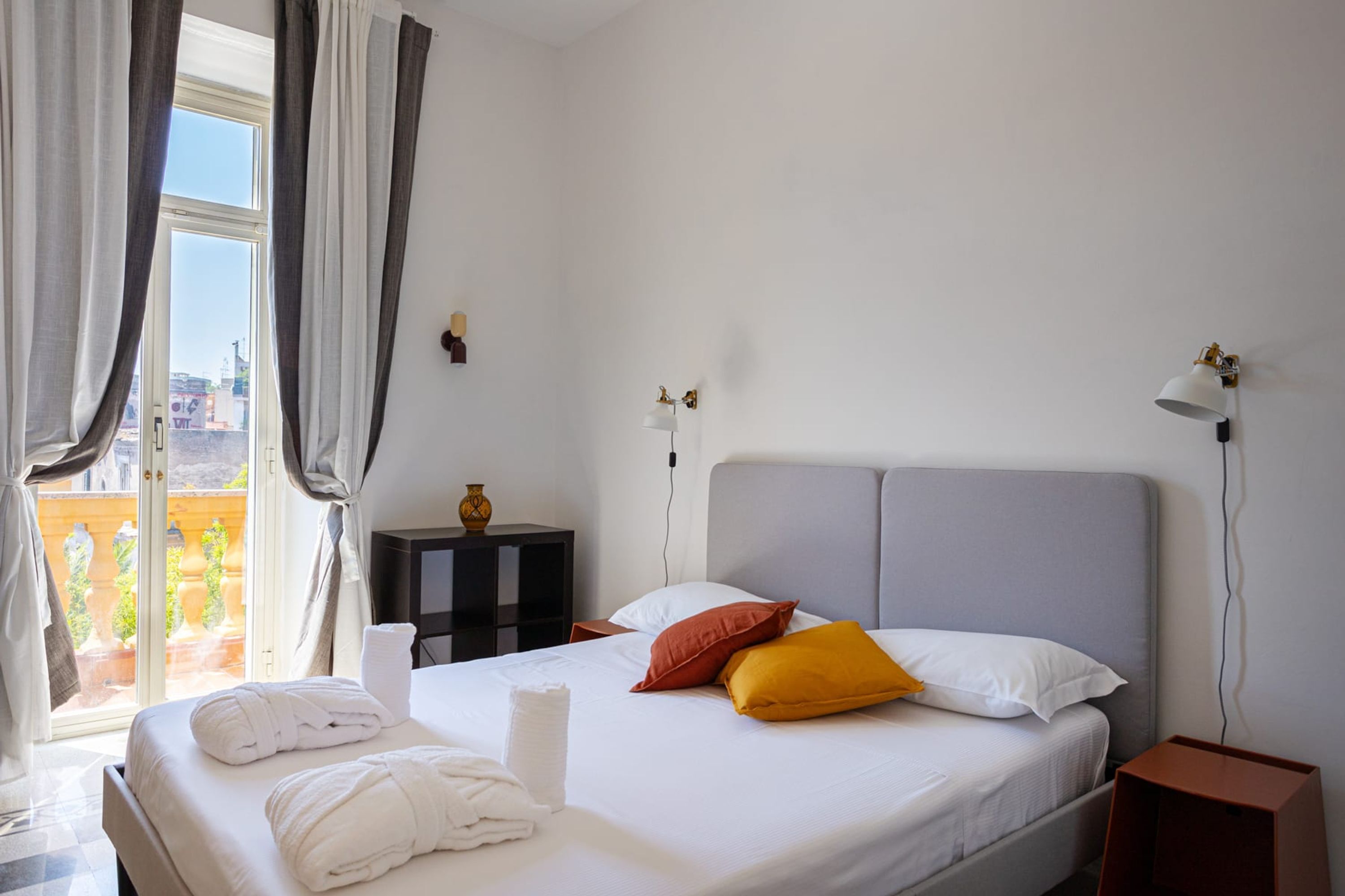A neatly made bed with white and yellow pillows, a bedside table with a lamp, and a view of the outdoors through a window with curtains.