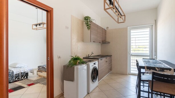 A compact kitchen with a washing machine, a dining table, and a view of the outside through a sliding door. - Cloud Swing Apartment by Wonderful Italy (Bari)