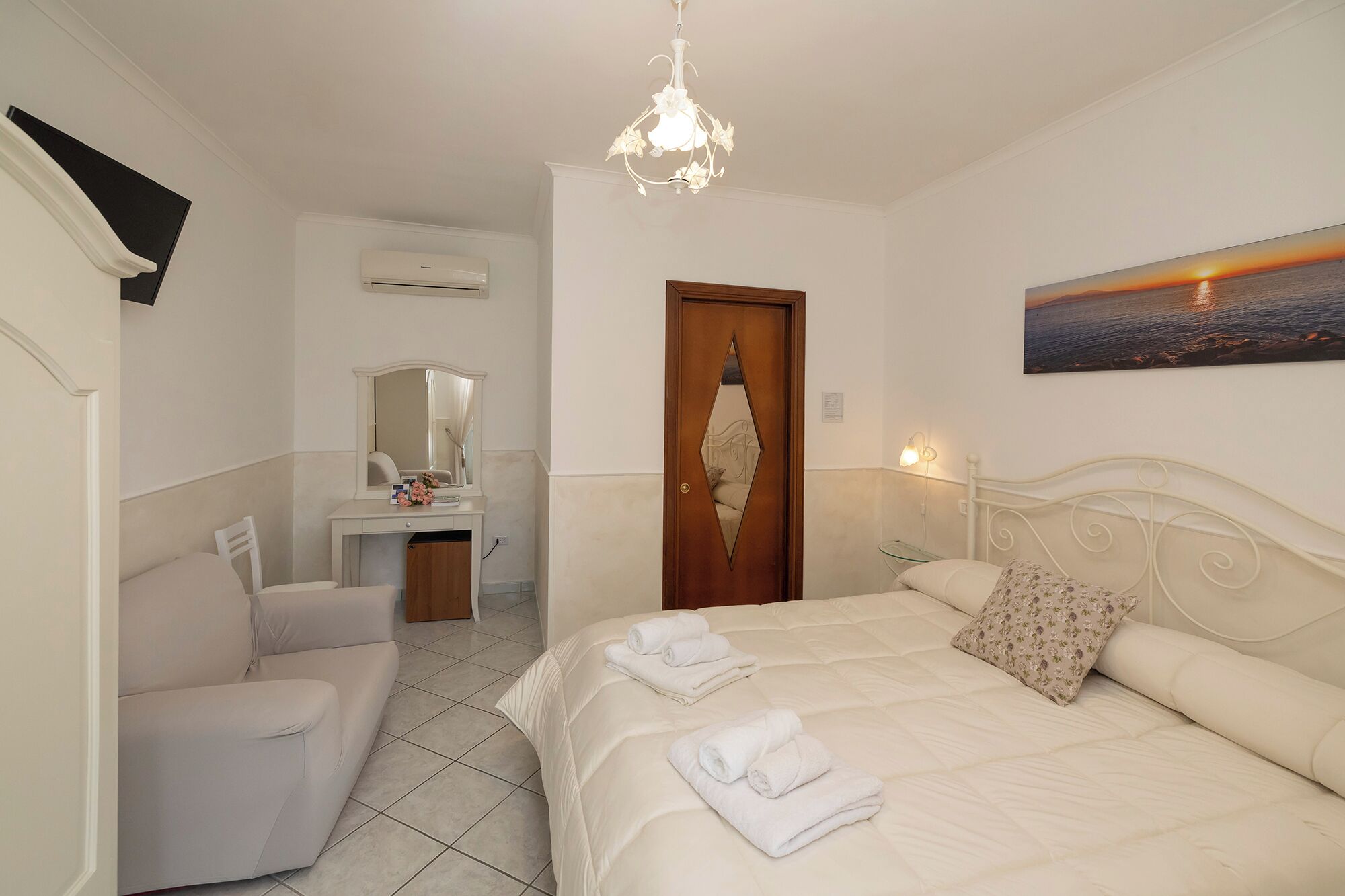 A bedroom with a bed, a desk with a mirror, a chair, a door, and a wall-mounted picture.