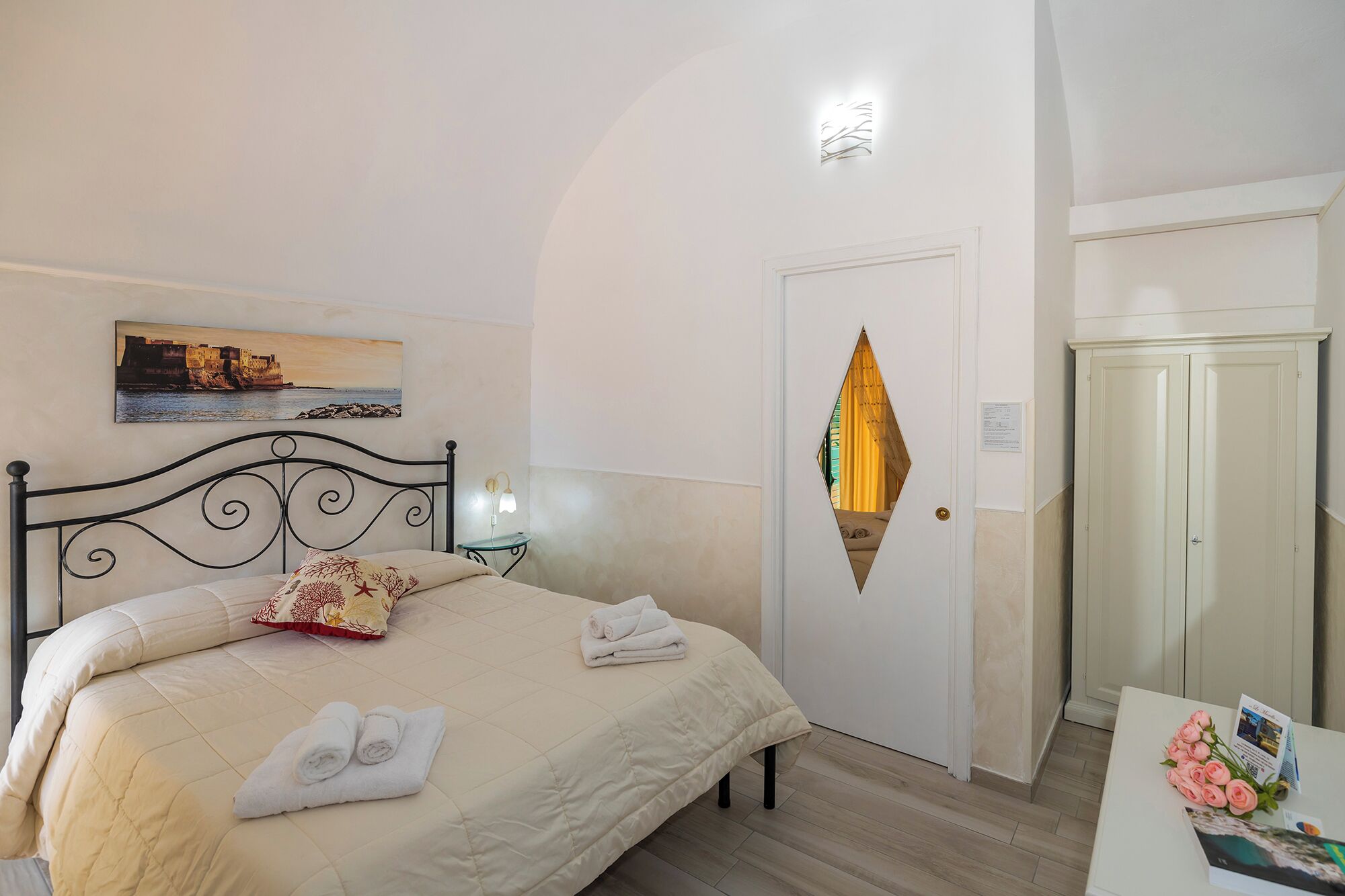 A bedroom with a bed, a nightstand, a wall-mounted light, a door with a decorative mirror, and a small table with a bouquet of flowers.