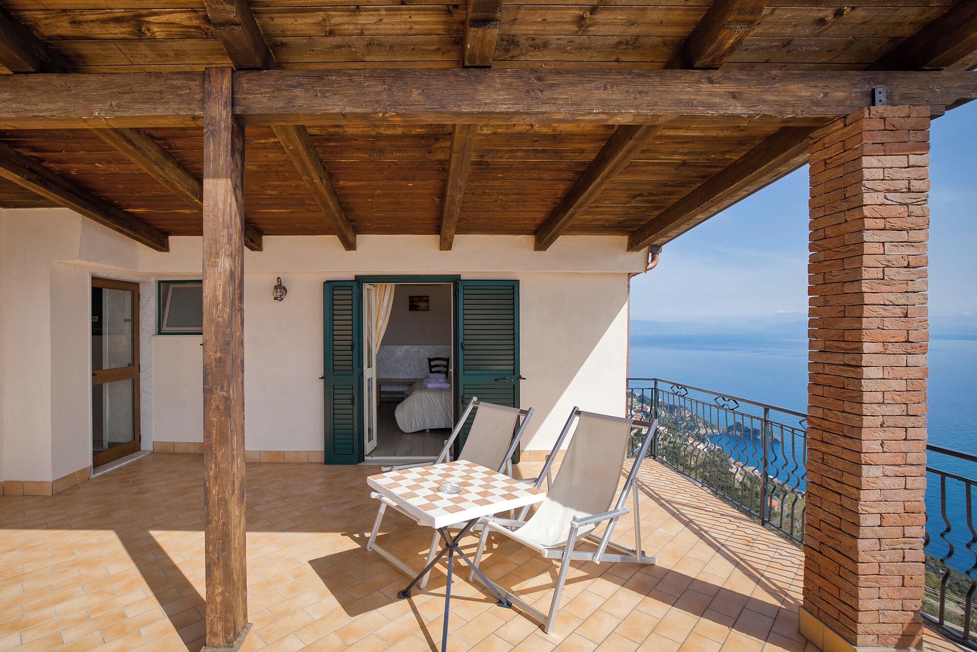 A balcony with a checkered table and chairs, a wooden ceiling, a brick wall, and a view of the sea.