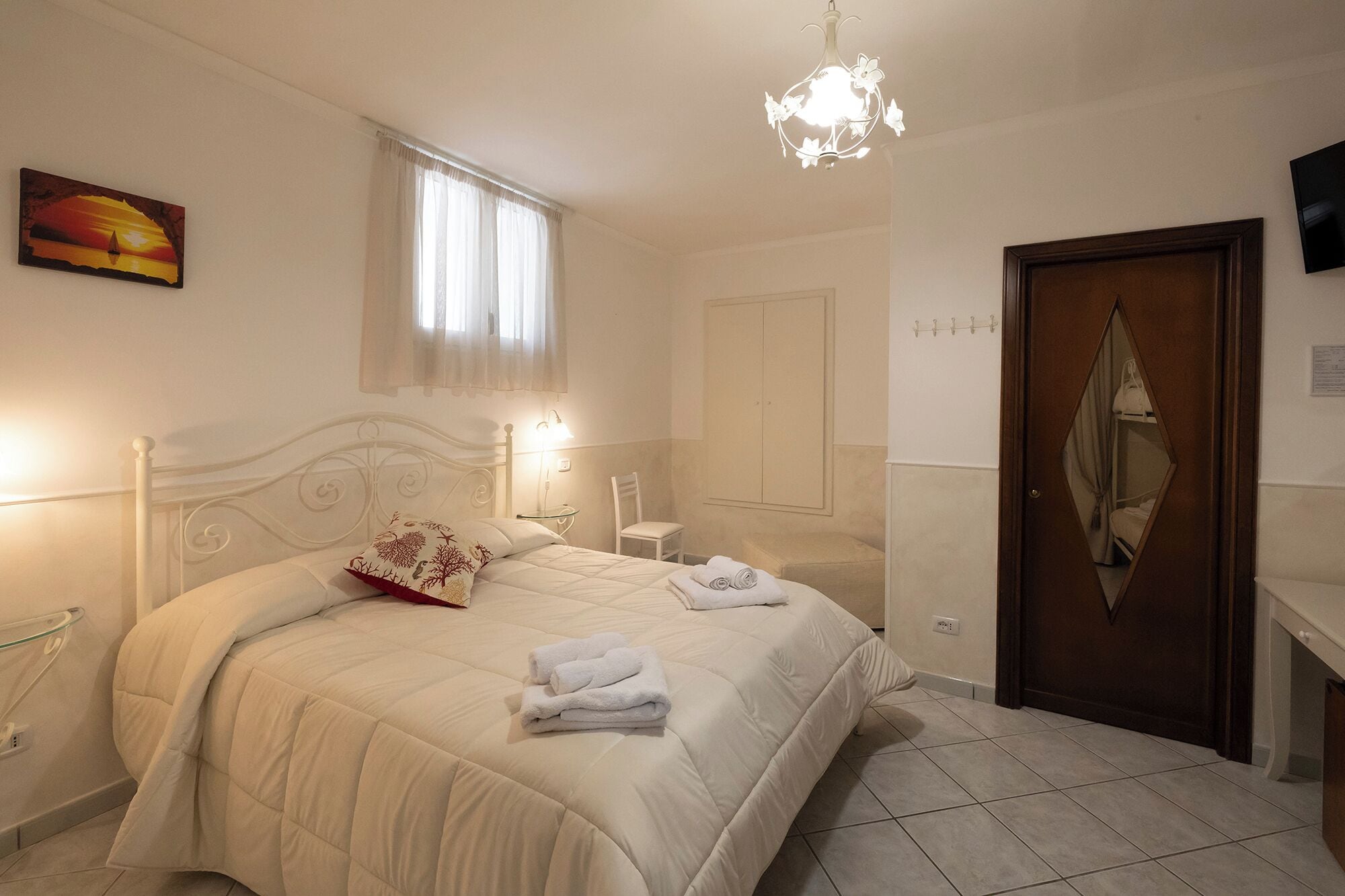 A well-lit bedroom with a large bed, a bedside table, a chair, a wardrobe, and a painting on the wall.