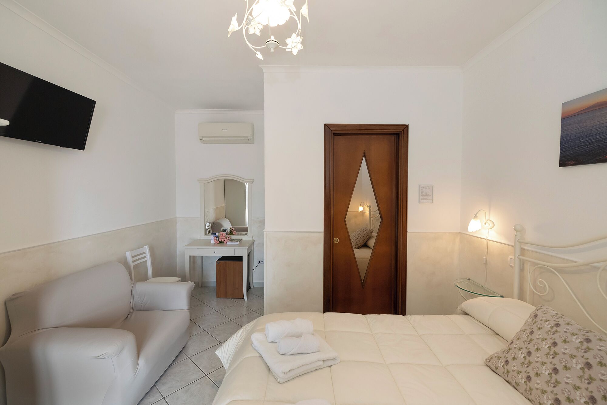 A hotel room with a bed, a sitting area with a sofa and a desk, a mirror, a wooden door with a mirror, and a painting on the wall.