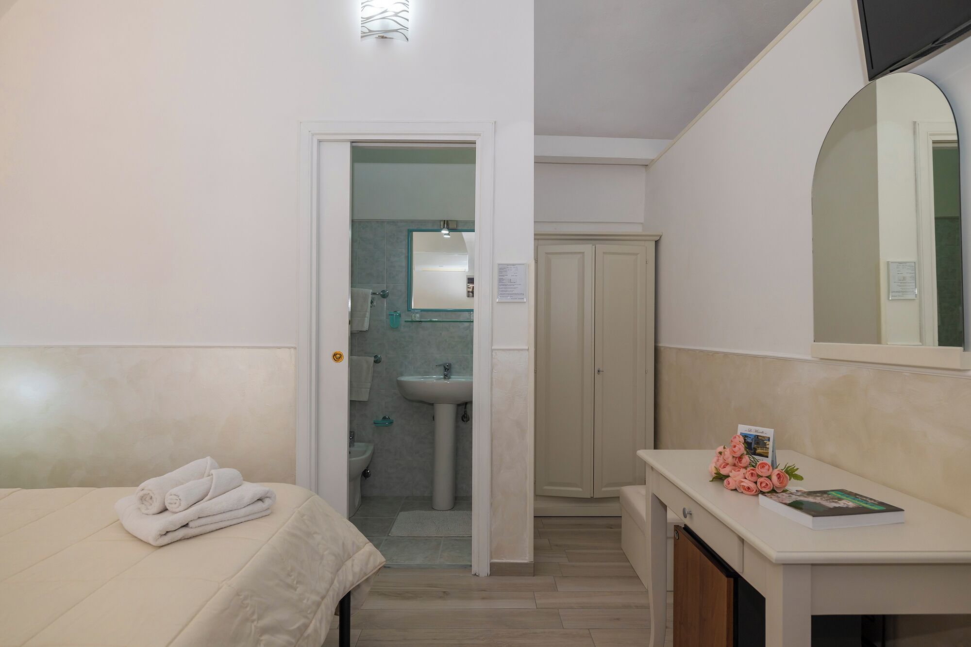 Camera d'albergo con un letto, un comodino con un libro e fiori, un bagno con lavandino e specchio, e un armadio.