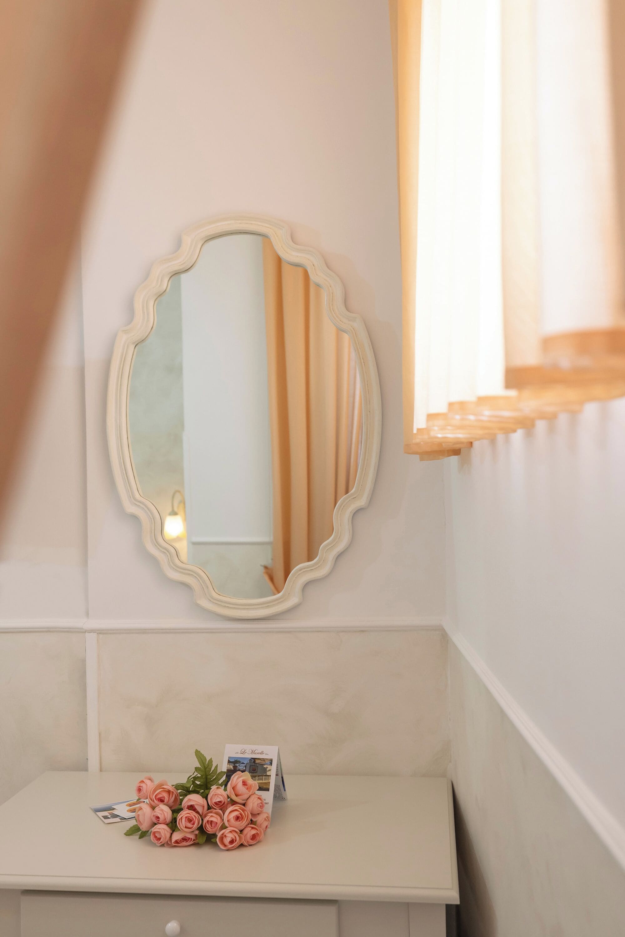 A bathroom with a white vanity, a round mirror with a wavy frame, and a bouquet of pink roses on the counter.