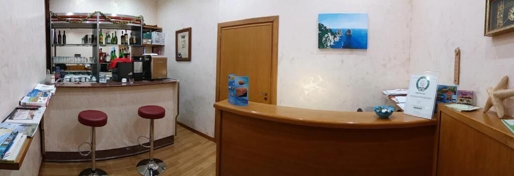 Un área de recepción con mostrador de madera, dos taburetes rojos, una máquina de café y una estantería con varios objetos.