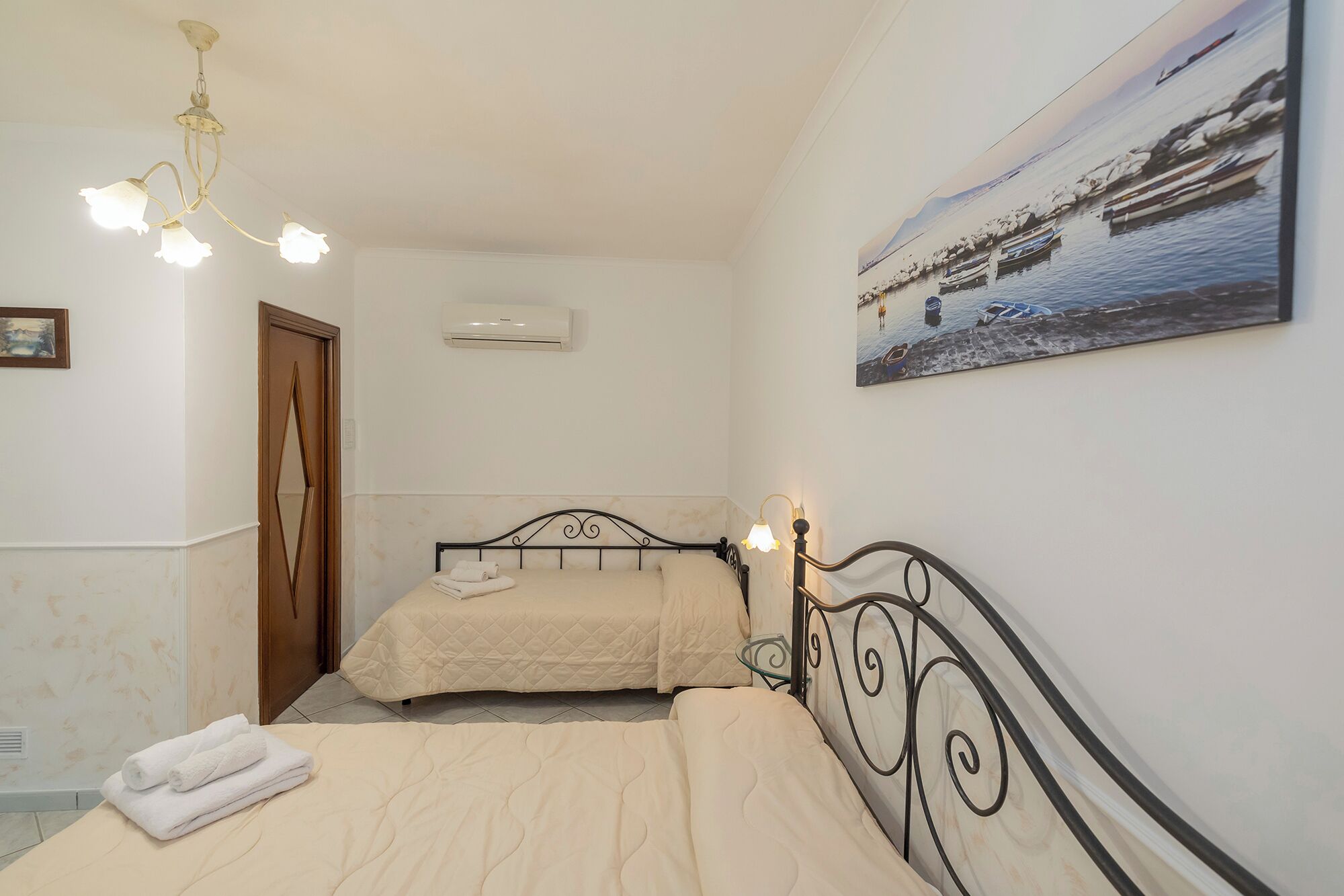 A bedroom with two beds, a chandelier, an air conditioner, a painting, and a door.