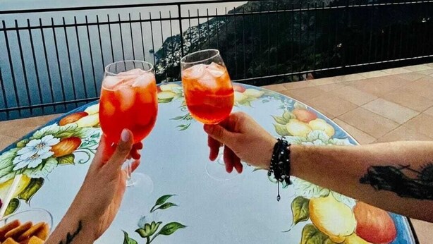Two people toasting with drinks on a table with a floral pattern, overlooking a scenic view of a sunset and mountains. - Le Marelle  (Furore)