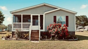 A single-story mobile home with a front porch, a small set of stairs leading to the entrance, and a garden with red flowers. - Clari's Place: 6mins to Holden Beach/Pets/Boat Parking/Plenty of Coastal Charm! (Supply)