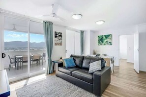 A modern living room with a black leather sofa, a dining area with a table and chairs, and a large sliding glass door leading to a balcony. - Cairns City Escape—Elevated Modern Haven with Pool (Cairns)