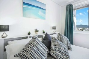 A hotel room with a bed, a painting of a beach, a window with a view of buildings, and a nightstand with a lamp and a plant.