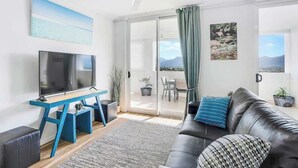 Ein modernes Wohnzimmer mit einem Flachbildfernseher, einem blauen Fernsehschrank, einem schwarzen Ledersofa, einem gemusterten Kissen, einem Deckenventilator und Blick ins Freie durch eine Glastür.