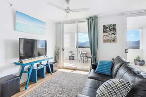 A modern living room with a flat-screen TV, a blue TV stand, a black leather sofa, a patterned cushion, a ceiling fan, and a view of the outdoors through a glass door.