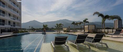 Zona de piscina con sillones reclinables, cielo despejado y montañas al fondo.