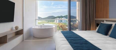 Habitación de hotel moderna con una cama grande, una bañera integrada y un balcón con vistas a la ciudad y las colinas.