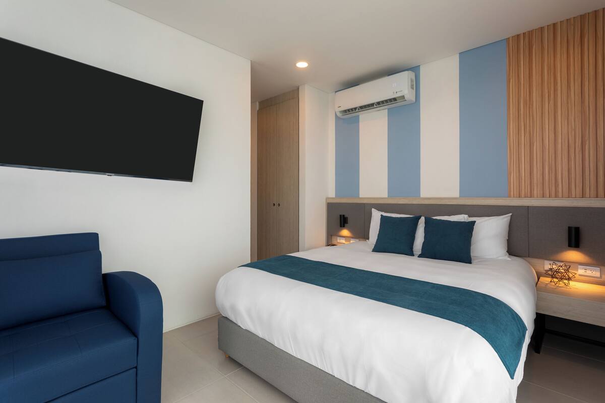 Une chambre d’hôtel moderne avec un grand lit, un fauteuil bleu, un téléviseur à écran plat et un climatiseur fixé au mur.