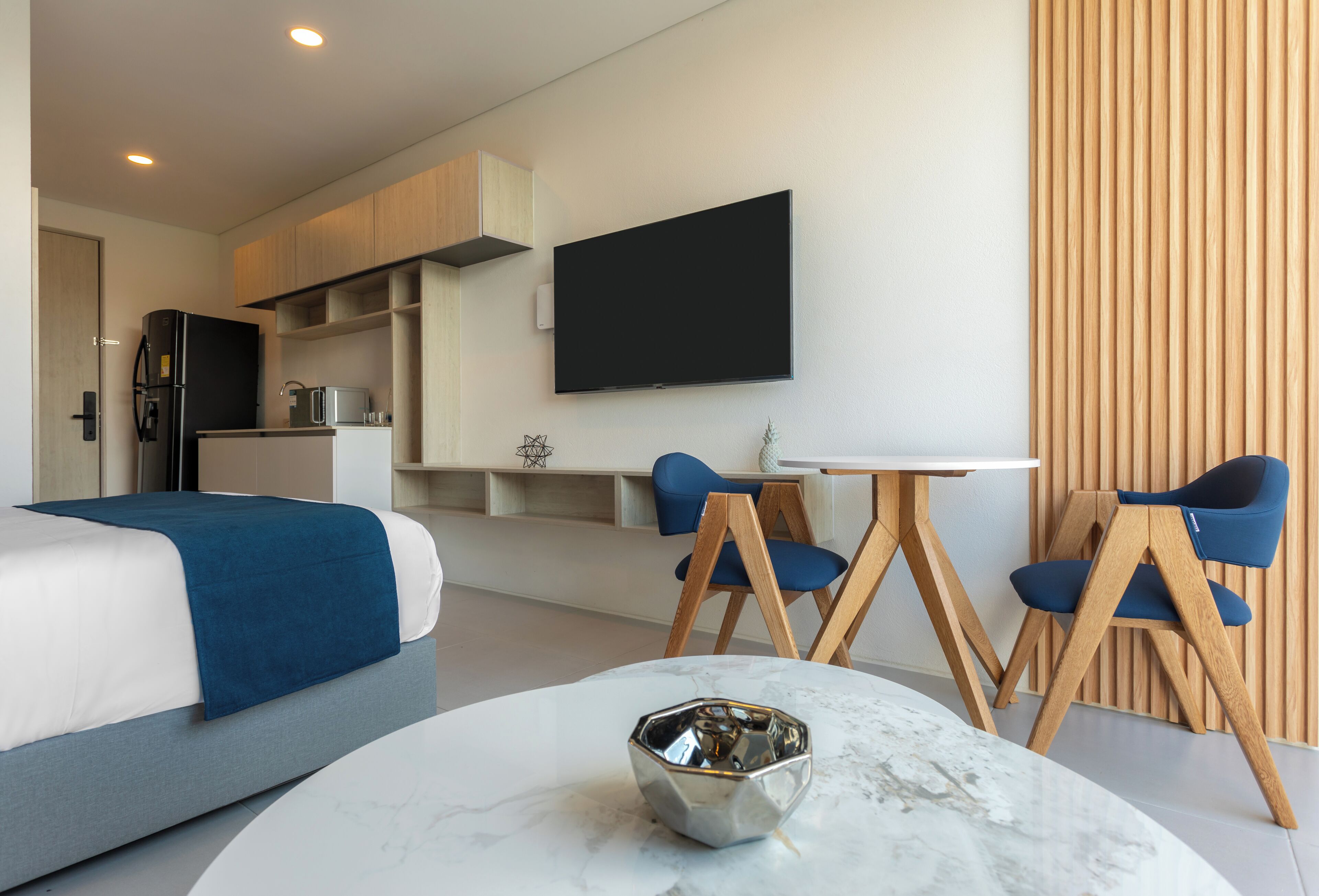 A modern hotel room with a bed, a dining table with chairs, a TV mounted on the wall, and a kitchen area.