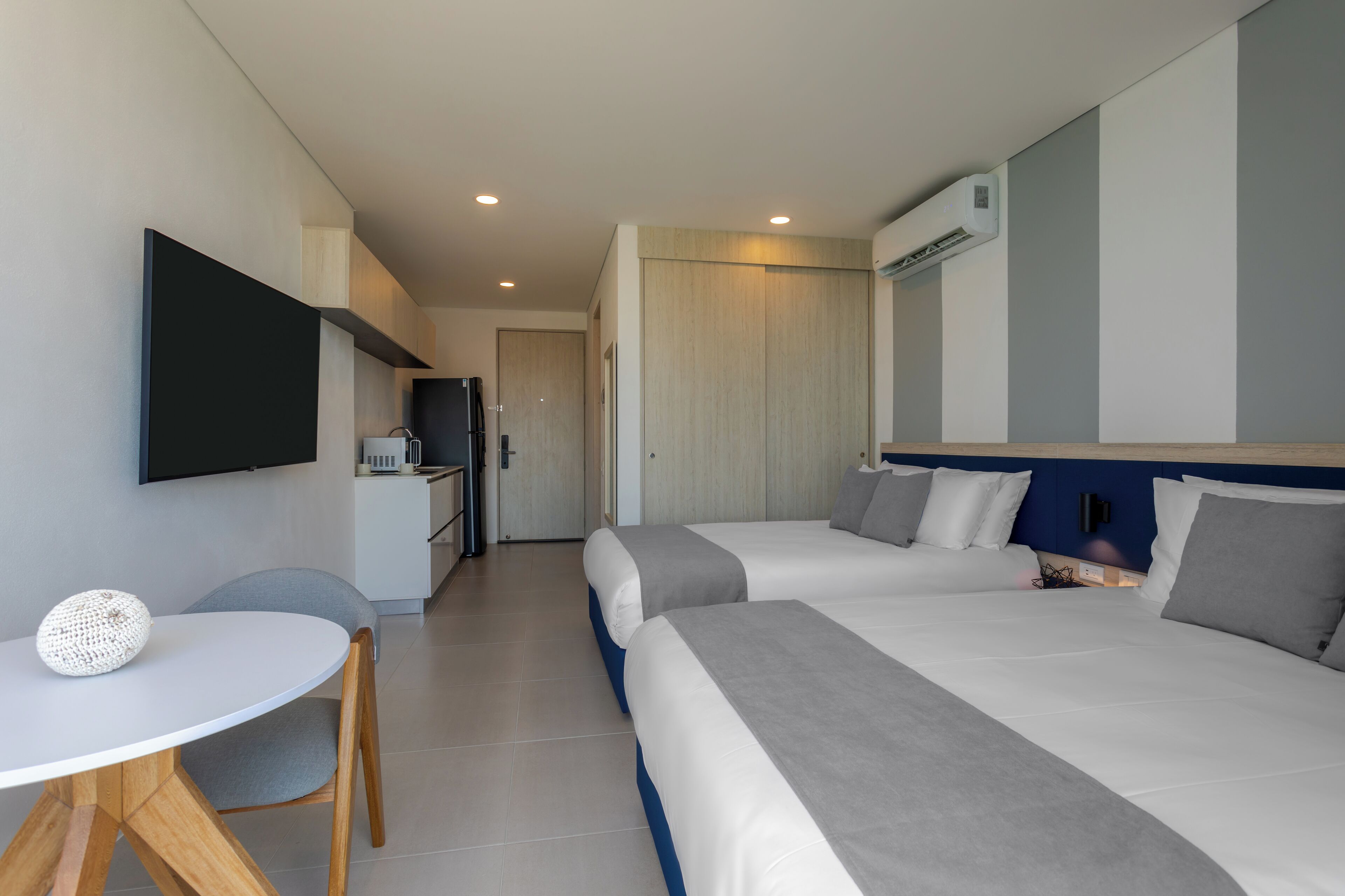 A modern hotel room with two beds, a small dining table, a TV, and a kitchenette.