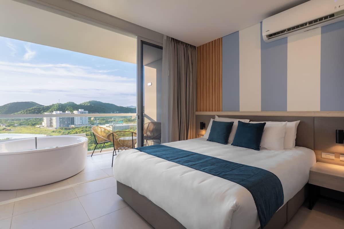 Une chambre d’hôtel moderne dotée d’un grand lit, d’une baignoire ronde et d’un balcon offrant une vue sur la ville et les collines.