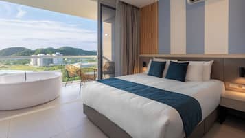 A modern hotel room with a large bed, a round bathtub, and a balcony with a view of the city and hills.