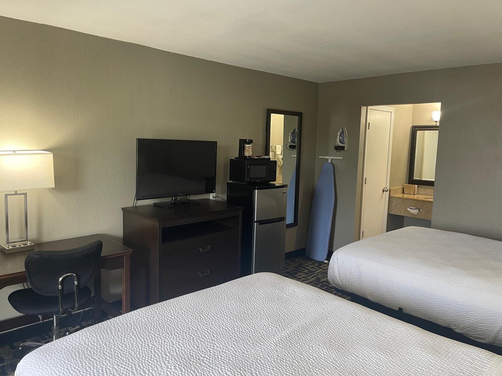 A hotel room with two beds, a desk with a TV, a microwave, an ironing board, and a bathroom with a mirror and sink.