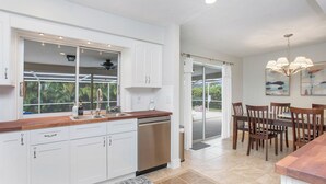 A modern kitchen with white cabinets, a wooden countertop, and a built-in dishwasher. There is a dining area with wooden chairs and a table, and a large window with a view of greenery outside.