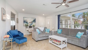 A spacious living room with a grey sofa set, a blue armchair, and a white coffee table. There is a ceiling fan, a large window with curtains, and a painting on the wall.