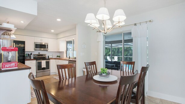 A modern kitchen with white cabinets, a microwave, and a popcorn machine. There is a dining area with a wooden table and chairs, and a large window offering a view of the outdoors.