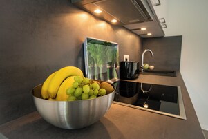 Una cocina con un tazón de acero inoxidable sobre la encimera, con plátanos y uvas, una cafetera negra y un espejo que refleja la cocina.