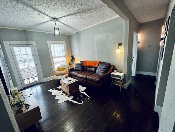 Un salon spacieux avec un canapé en cuir marron, une table basse blanche et une table d’appoint en bois. Il y a une grande fenêtre avec des stores, un ventilateur de plafond et un luminaire fixé au mur.
