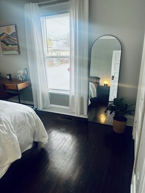 Une chambre à coucher avec un lit, un bureau doté d’un miroir, une plante en pot et une fenêtre avec des rideaux.
