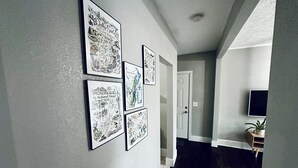 Un pasillo decorado con mapas y ilustraciones enmarcados en la pared, que conduce a una puerta y un cuarto con televisión y planta.