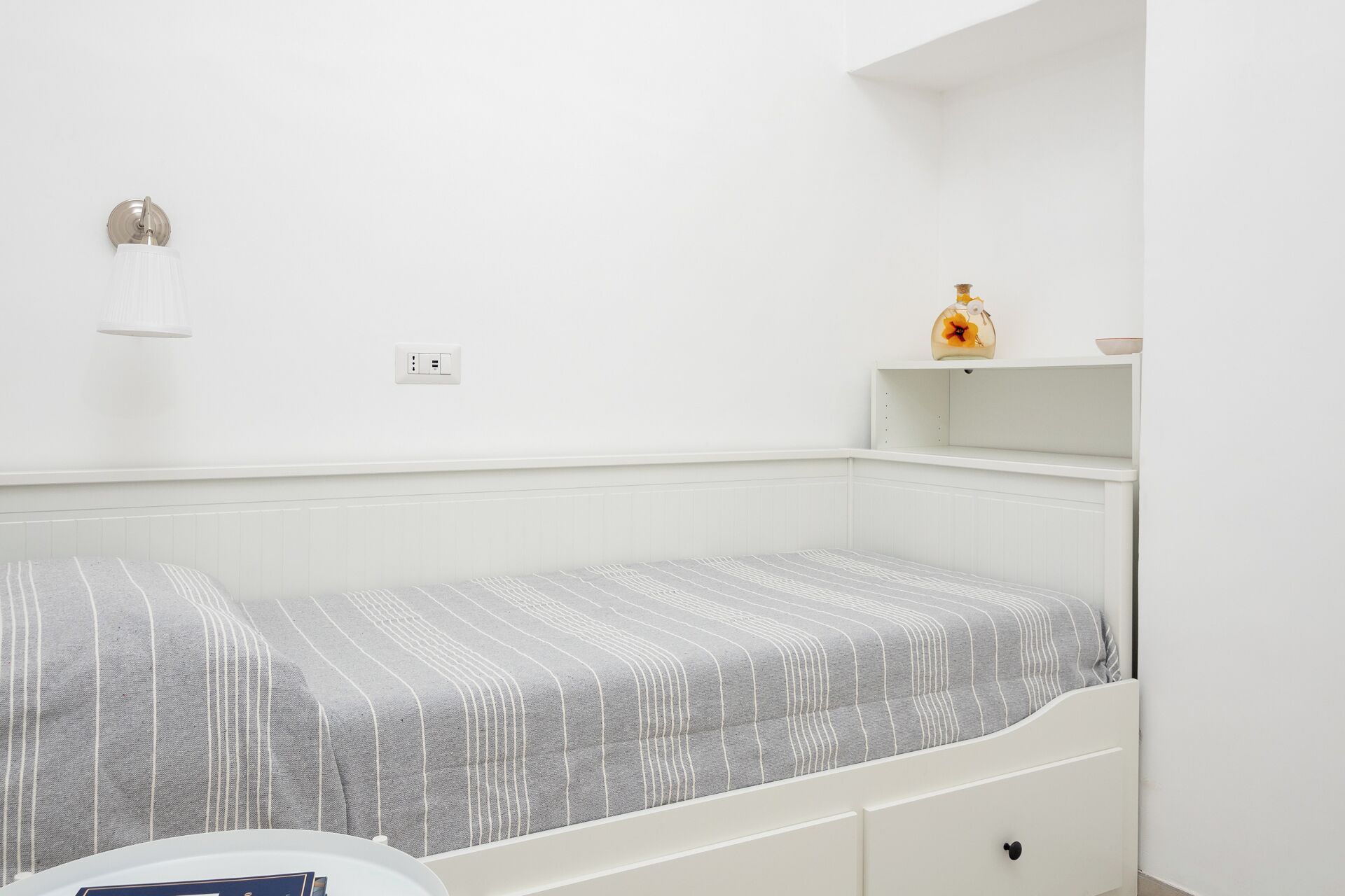 A small, white, single-bed room with a striped bedspread, a bedside table, a shelf with a vase, and a wall-mounted light fixture.