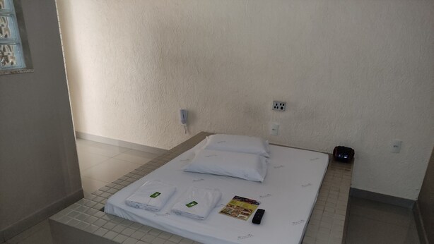 A neatly made bed with white linens and pillows, a remote control, and a brochure on the bed. - Motel Classe A  (Limeira)