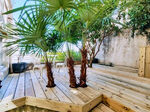 Een houten terras met potpalmen, witte stoelen en een betonnen muur op de achtergrond.