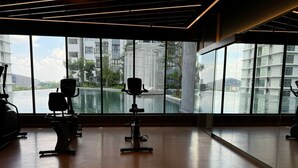Een fitnessruimte met trainingsapparatuur en grote ramen met uitzicht op een waterpartij en gebouwen.